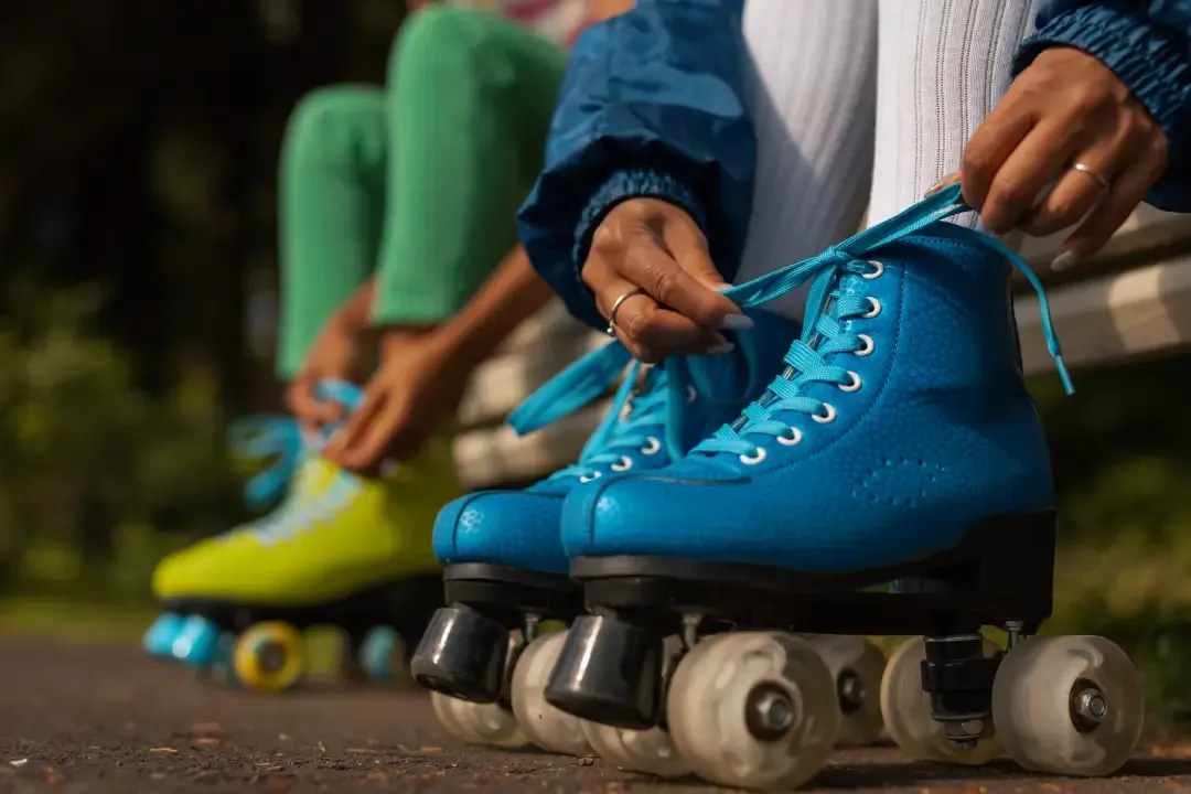 Nahaufnahme: Eine Person schnürt blaue Rollschuhe auf einem Parkweg, im Hintergrund eine zweite Person mit gelben Rollschuhen